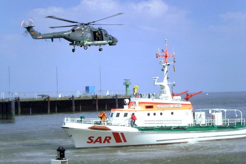 image of a gray marine helicopter hovering over a white search and rescue ship in the harbor